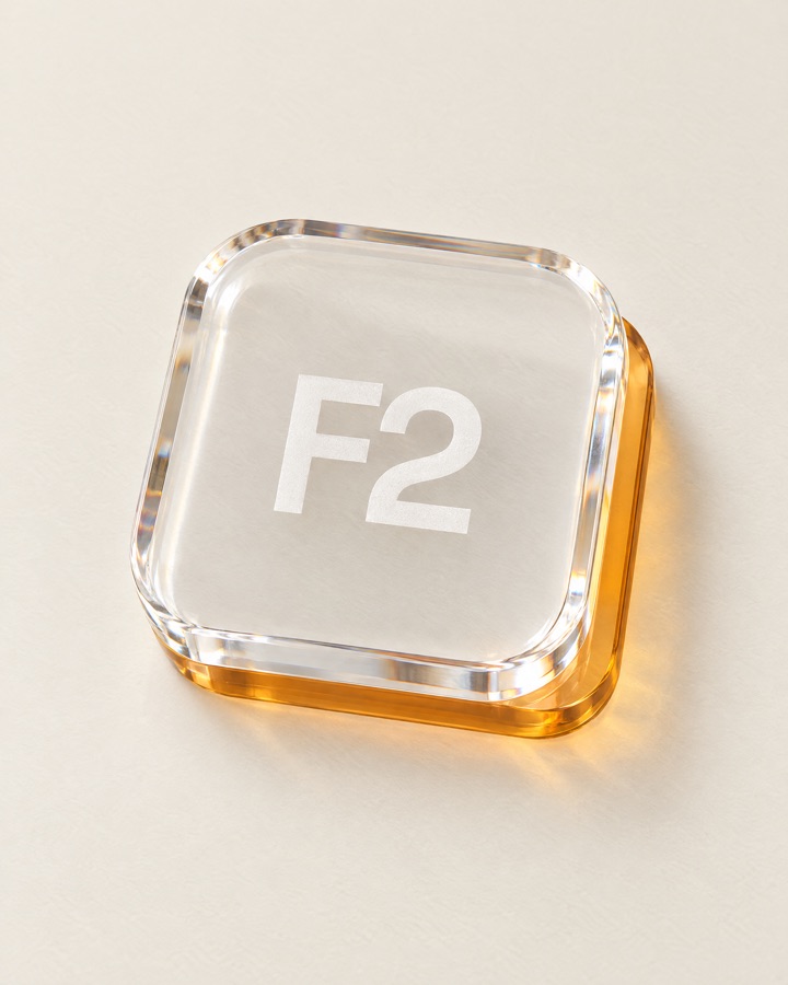 F2 keycap rendered in clear optical crystal over a solid amber edge, lying on a bone surface